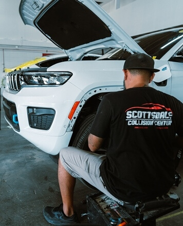 A technician inspecting the front of a white SUV inside an auto body repair shop