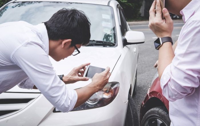 Two men gets caught in a fender bender collision. One leans over to take a photo of the damage and the other calls the authorities