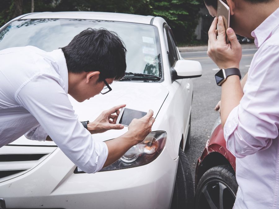 Two men get caught in a fender bender collision. One leans over to take a photo of the damage, and the other calls the authorities