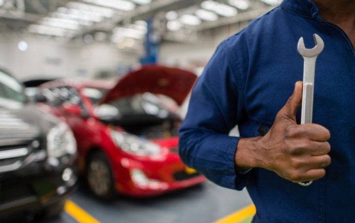 A close-up of a mechanic in blue coveralls holding a silver wrench, with blurred cars and an open hood in the background