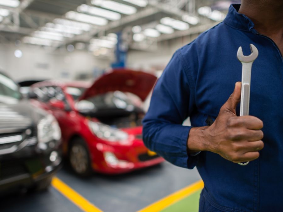 A close-up of a mechanic in blue coveralls holding a silver wrench, with blurred cars and an open hood in the background