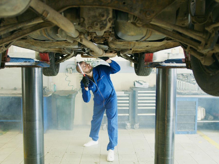 A mechanic in blue coveralls uses a flashlight to inspect the complex underside of a car raised high on a hydraulic lift